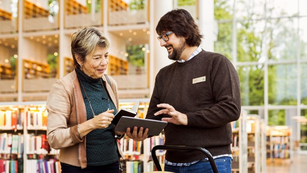 En äldre dam står i samtal med en yngre bibliotekarie i ett stort ljust rum. Bilden är länkad till digiteket.se.