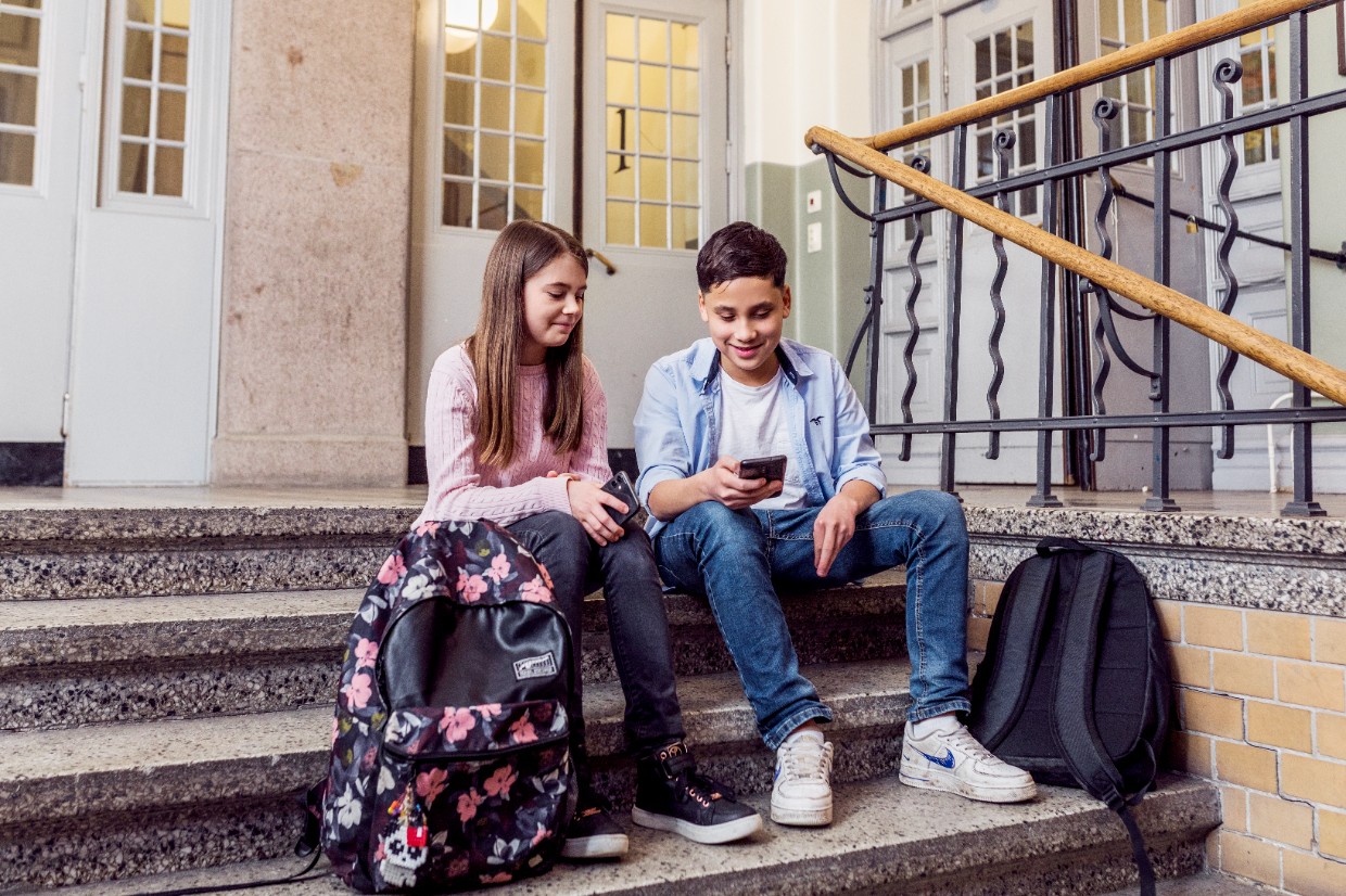 Två mellanstadieelever sitter i en trappa i en skola. De tittar på sina mobiler och ler. Vid fötterna har de sina ryggsäckar.