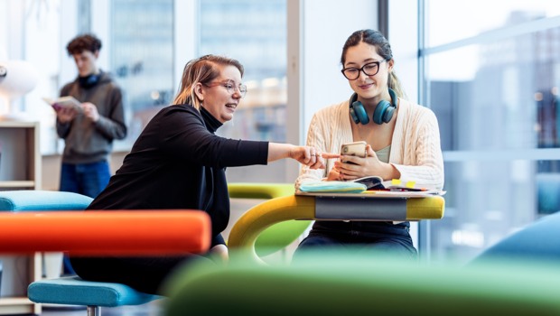 En bibliotekarie och en student sitter vid en studieplats i biblioteket. Bibliotekarien lutar sig fram och pekar på studentens mobil som denne håller i handen. Studenten har hörlurar runt halsen och på bordet ligger hennes kurslitteratur.