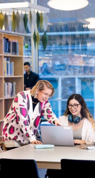 En bibliotekarie står bredvid en student vid ett bord i högskolebiblioteket. Bibliotekarien lutar sig fram och pekar på skärmen. På bordet ligger kurslitteratur och studenten har hörlurar hängande runt halsen.