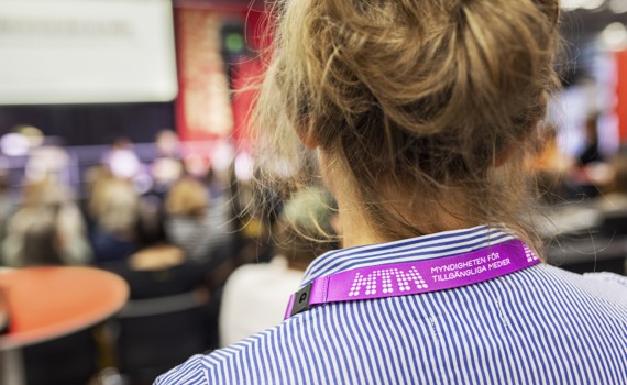 En kvinna står i en seminariesal, hon har ett nyckelband runt halsen där det står MTM