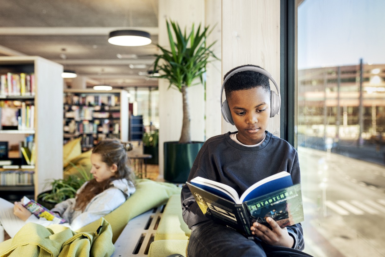 En mellanstadieelev sitter vid ett fönster i ett skolbibliotek och läser en pappersbok. Han har stora hörlurar på sig. Han tittar koncentrerat ner i boken. Bakom honom sitter en tjej i samma ålder och läser. Det skymtar bokhyllor och växter i bakgrunden.