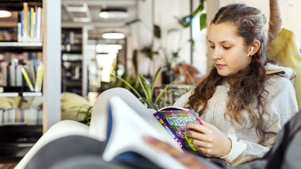 En flicka sitter och läser en bok och lyssnar samtidigt i hörlurar.