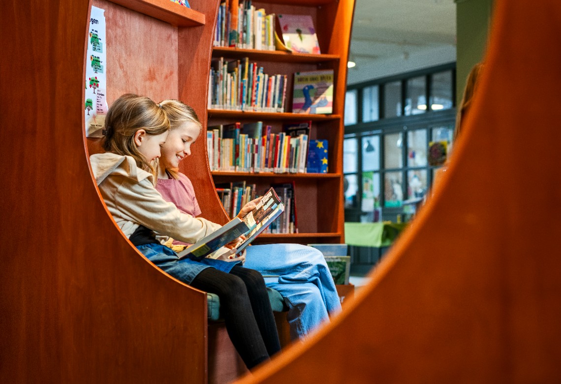 Två flickor sitter i en mörkröd sittgrupp på ett bibliotek och läser tillsammans i en bilderbok. De ler.