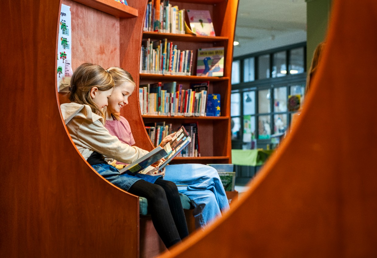 Två barn sitter i en soffa i ett bibliotek. De håller i varsin uppslagen bok. Det ena barnet pekar på något i den andras bok och de skrattar.