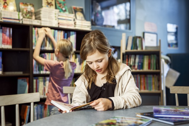 Ett barn sitter vid ett bord i ett bibliotek och läser en tryckt bok. På bordet ligger flera andra böcker utspridda. I bakgrunden plockar ett barn med böcker i en bokhylla.