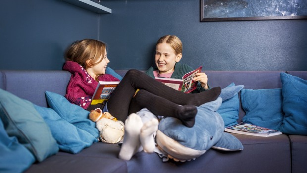  Två barn sitter uppkrupna i en soffa i ett bibliotek med varsin uppslagen bok i händerna. De tittar på varandra och skrattar.