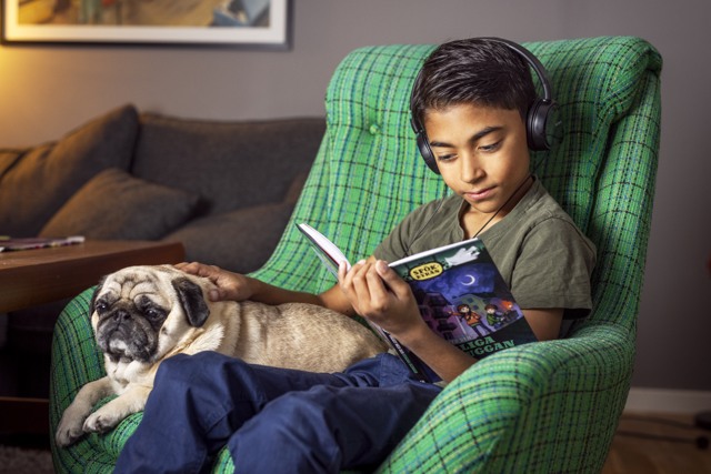  Ett barn sitter i en fåtölj tillsammans med en liten hund och läser en bok. Han håller i den tryckta boken med ena handen, med den andra handen klappar han hunden. På huvudet har han svarta hörlurar.