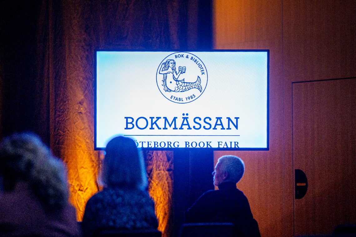 Ett foto av en LED-skärm där det står Bokmässan. Nedanför skärmen syns tre personer som sitter och lyssnar. Foto:Apelöga/MTM