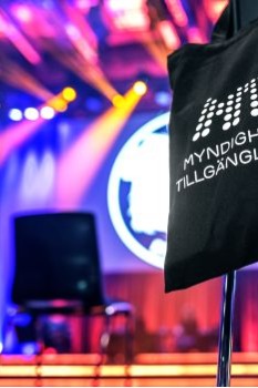 Fotot visar MTMs svarta tygkasse med vit logotyp i en färggrann miljö, ett rum med många strålkastare och man anar en stol framför en scen.
